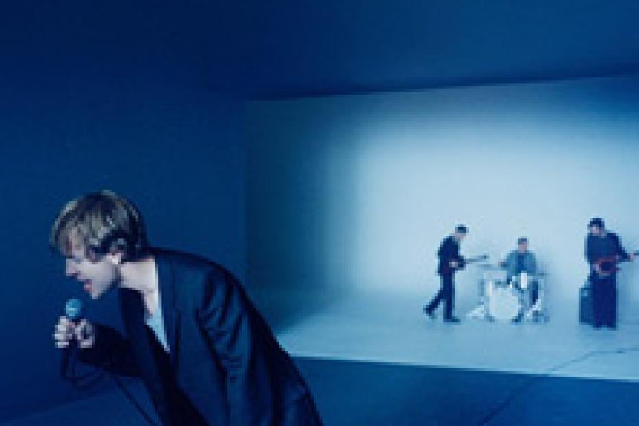 Eine Band spielt in einem blauen Raum, wobei der Sänger im Vordergrund steht.