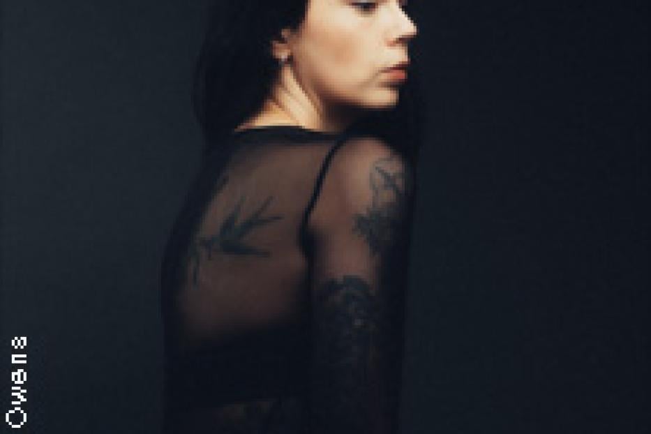 Eine Frau mit dunklen Haaren und Tätowierungen blickt über ihre Schulter.