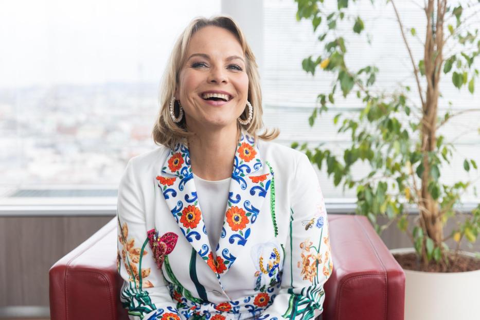 Eine lachende Frau, vermutlich Elina Garanca, sitzt in einem floralen Blazer.