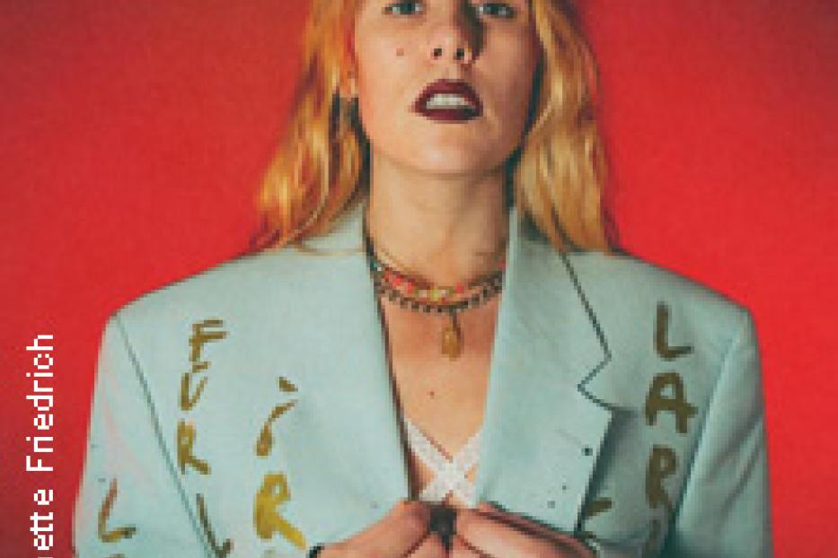 Eine Frau mit blonden Haaren und einem hellblauen Blazer vor einem roten Hintergrund.