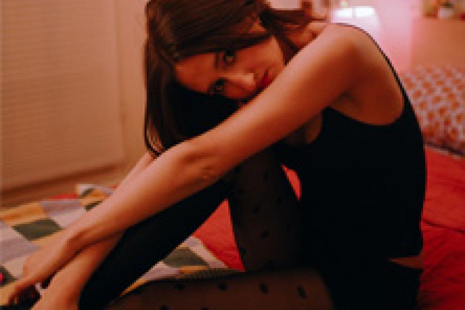 Eine junge Frau sitzt auf einem Bett und umarmt ihre Knie.