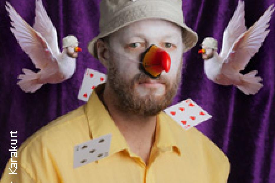Ein Mann mit Clownsschminke, Hut und roter Nase posiert mit Spielkarten und Tauben vor einem lila Hintergrund.