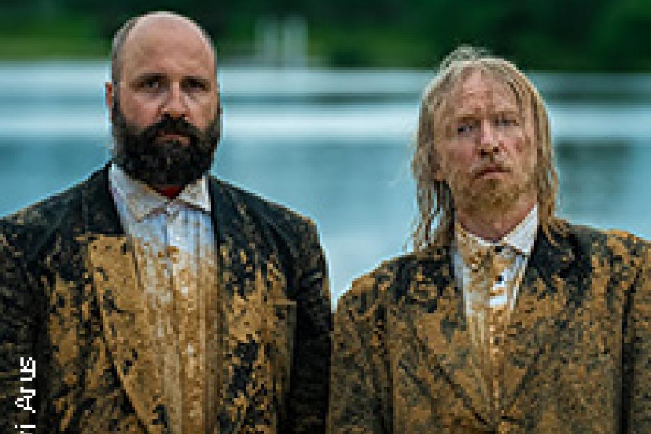 Zwei Männer in schlammverschmierten Anzügen stehen vor einem See.