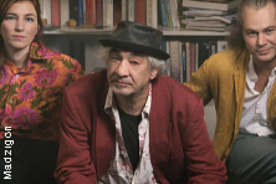 Drei Personen sitzen vor einem Bücherregal, der Mann in der Mitte trägt einen Hut.