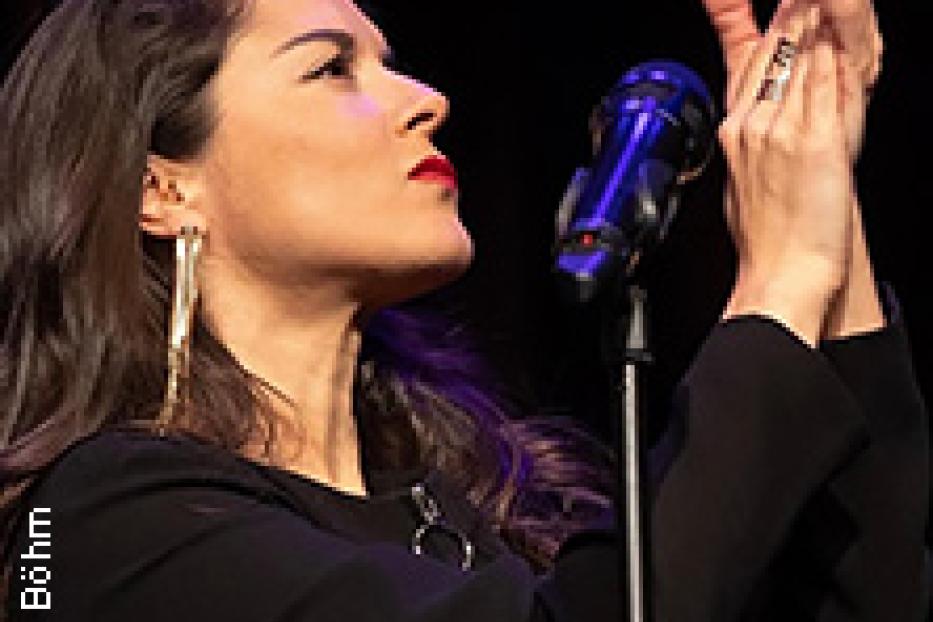 Eine Sängerin mit roten Lippen und langen Ohrringen steht am Mikrofon und klatscht in die Hände.