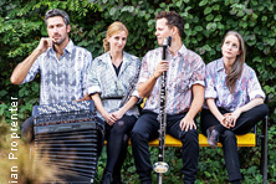 Eine vierköpfige Band sitzt mit Akkordeon und Klarinette auf einer Bank vor einer grünen Hecke.