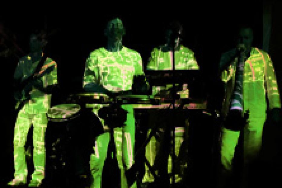 Eine Band tritt in fluoreszierender Kleidung auf einer dunklen Bühne auf.