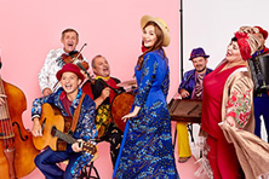 Eine Gruppe von Musikern in farbenfrohen Kostümen spielt auf verschiedenen Instrumenten vor einem rosa Hintergrund.