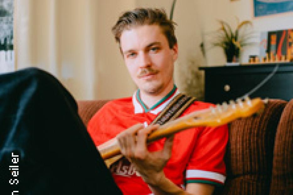 Ein Mann mit Schnurrbart sitzt auf einem Sofa und hält eine E-Gitarre.
