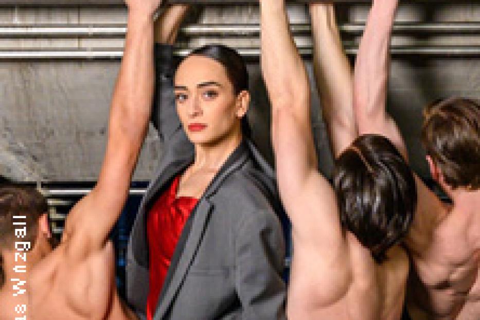 Eine Frau in einem roten Kleid und einem grauen Blazer steht umgeben von mehreren Männern, die sich an Metallstangen festhalten.