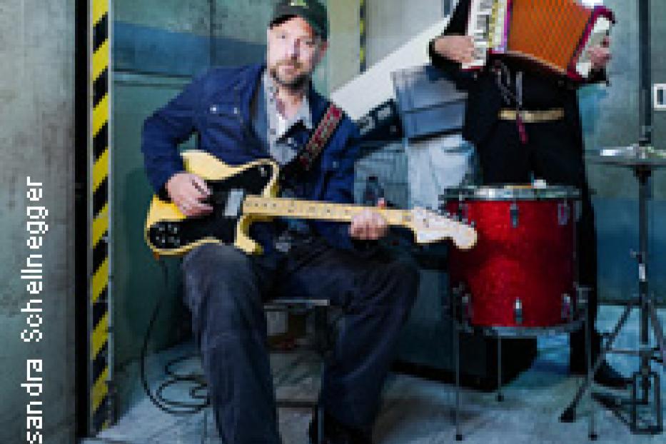 Zwei Musiker spielen Gitarre und Akkordeon vor einem roten Schlagzeug.