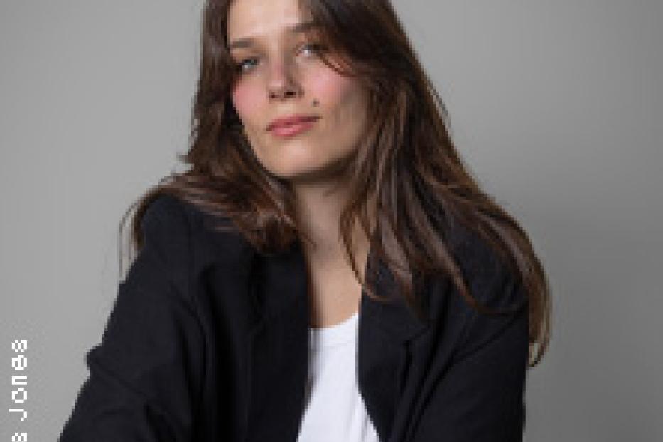 Eine junge Frau mit braunem Haar und einem schwarzen Blazer vor einem grauen Hintergrund.