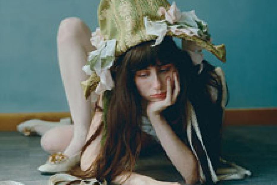 Eine junge Frau mit langen, dunklen Haaren und einem großen, verzierten Hut posiert auf dem Boden.