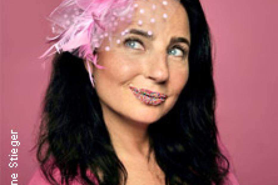 Eine Frau mit dunklen Haaren und einem rosa Hut mit einem Lolli vor einem rosa Hintergrund.