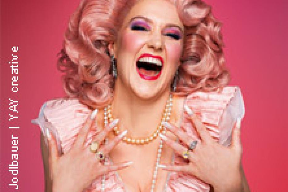 Eine Dragqueen mit rosa Perücke, üppigem Make-up und einem rosa Kleid lacht.