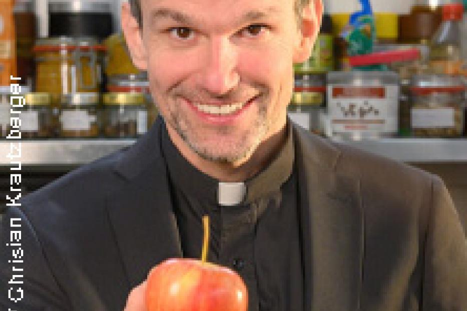 Ein lächelnder Priester hält einen roten Apfel in der Hand.