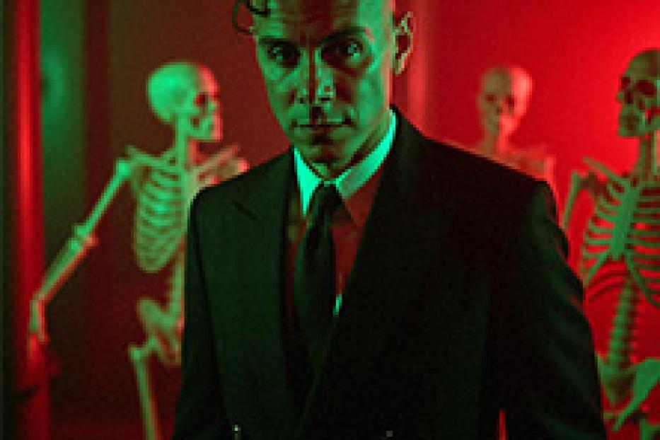 Ein Mann in einem Anzug steht vor drei Skeletten, beleuchtet in Rot und Grün.