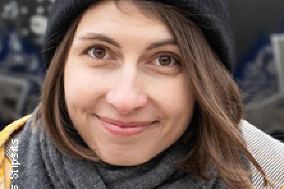 Eine Frau mit braunen Haaren, Mütze und Schal lächelt in die Kamera.