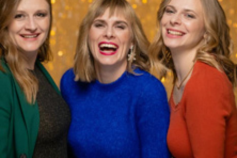 Drei Frauen stehen vor einem goldenen Hintergrund und lachen.