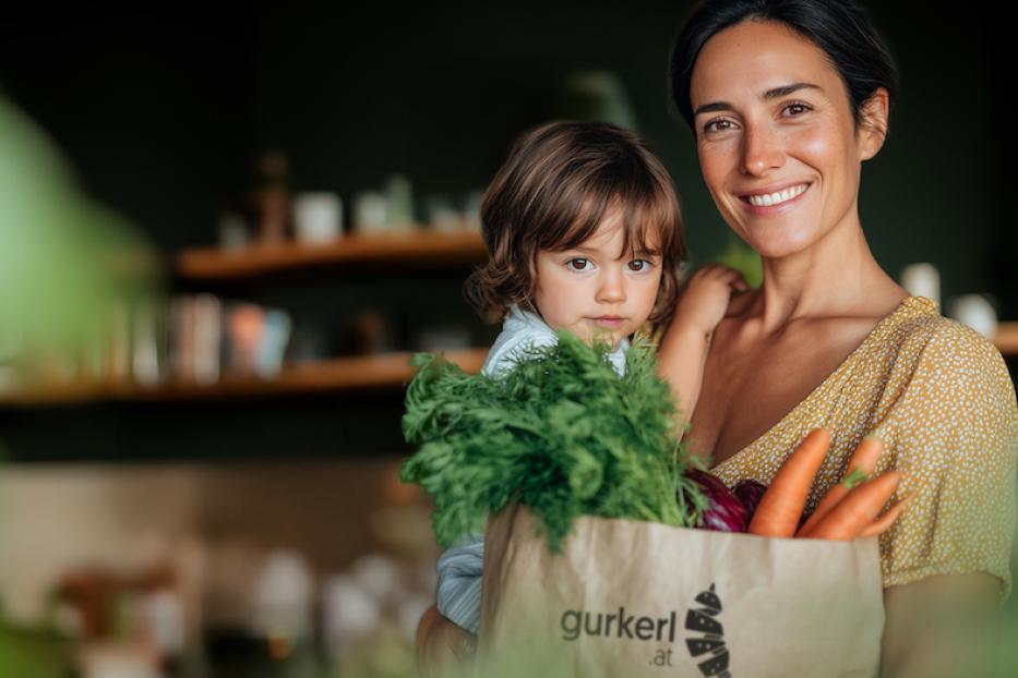 Eine Frau hat ihr Kind auf ihrem Arm und freut sich über ihre Einkaufstüte von gurkerl. 
