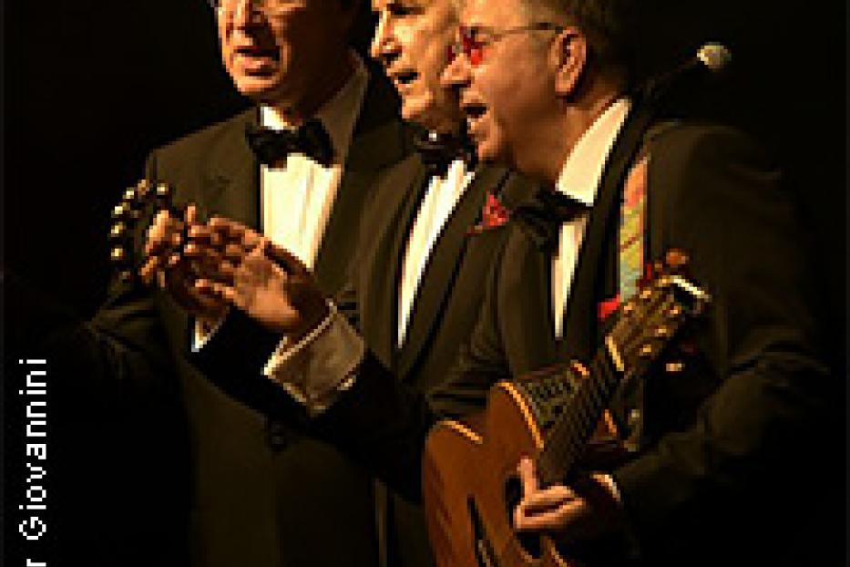 Drei Männer in Anzügen singen und spielen Gitarre auf einer Bühne.