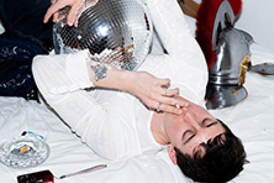 Ein Mann liegt mit einer Discokugel und einem römischen Helm auf einem Bett und raucht.