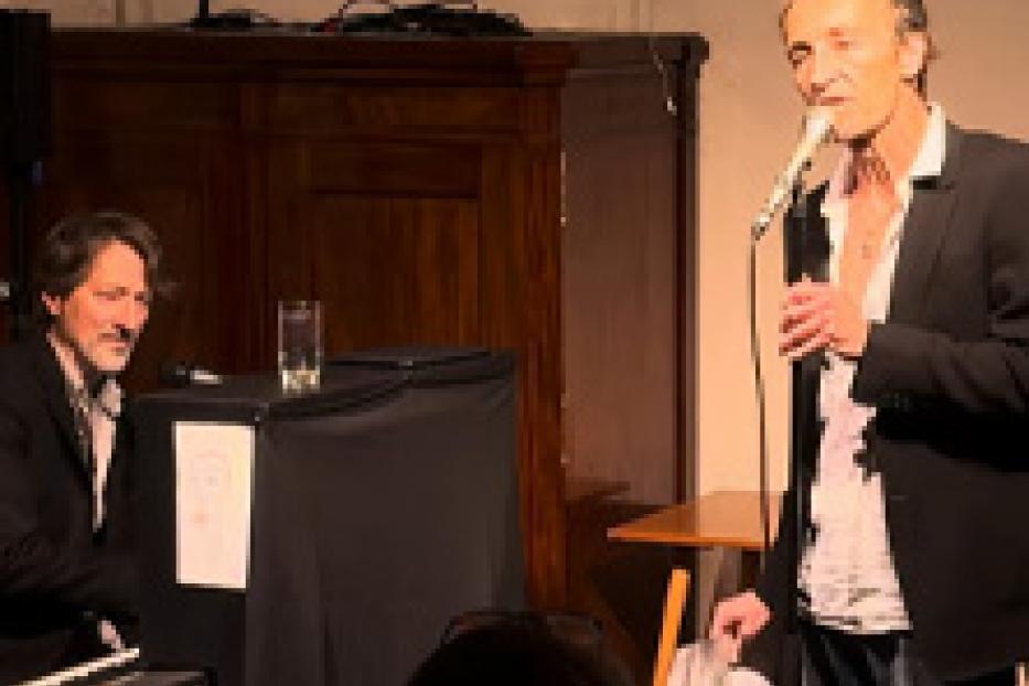 Ein Mann singt mit Mikrofon, während ein anderer Klavier spielt.