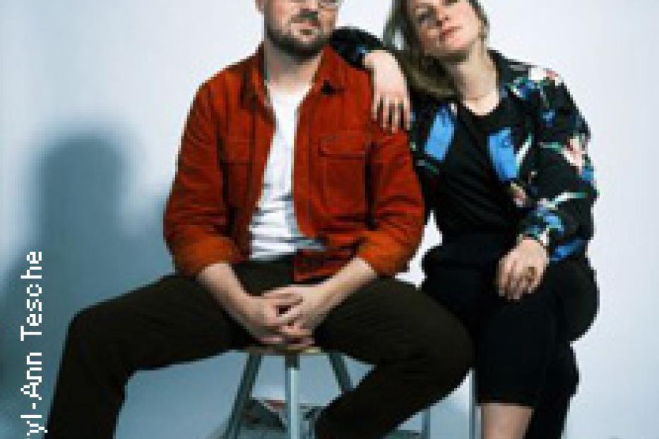 Ein Mann und eine Frau posieren auf einem Hocker vor einer weißen Wand.