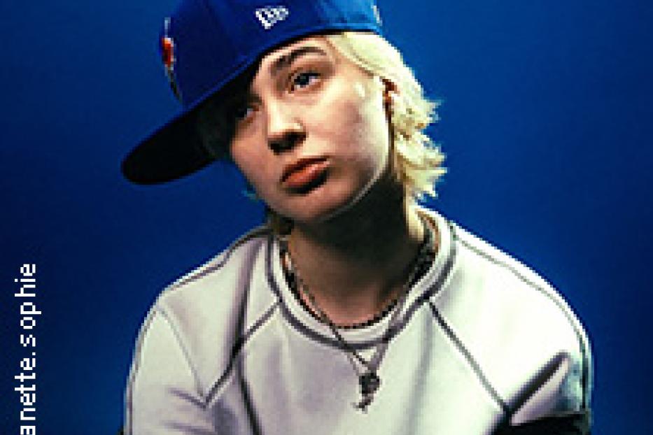 Eine junge Frau mit kurzer, blonder Haare und einer blauen Baseballkappe posiert vor einem blauen Hintergrund.