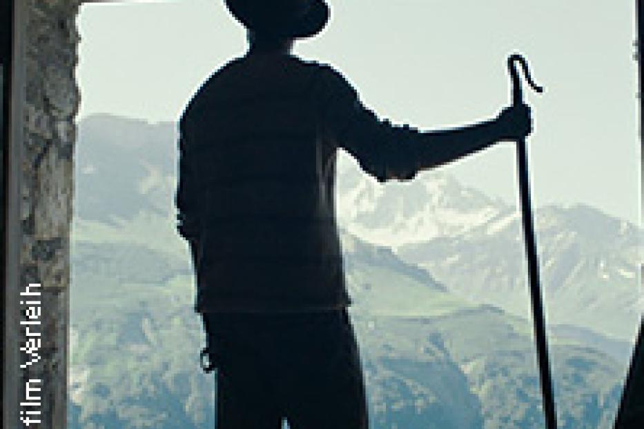 Ein Mann mit Hut und Stock blickt aus einer Hütte auf die Berge.
