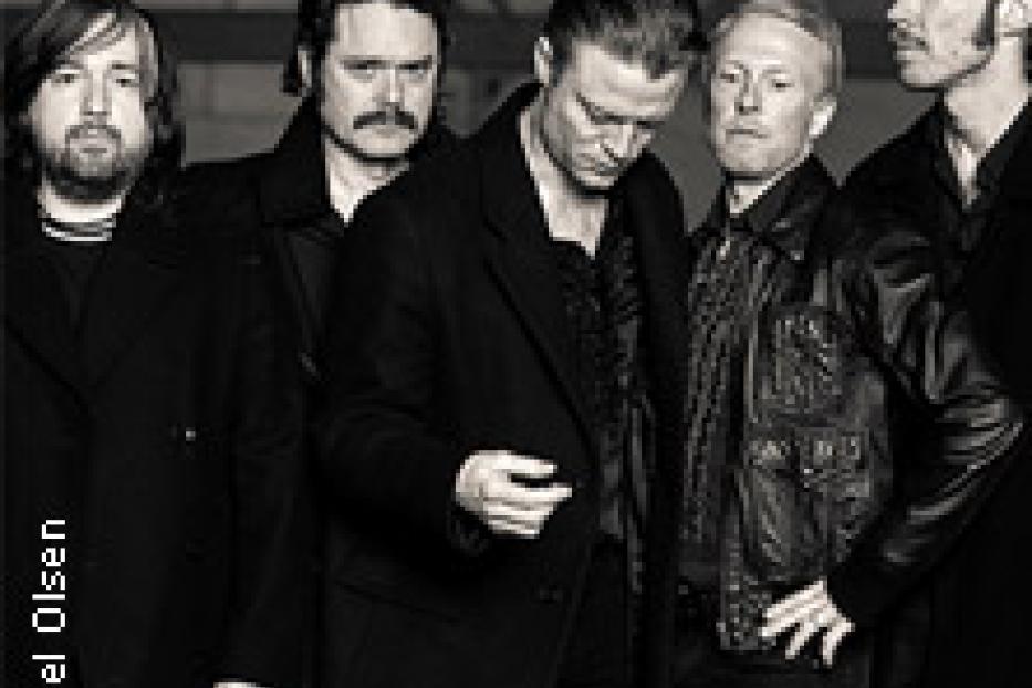Die Band Interpol steht in dunkler Kleidung vor einer grauen Wand.