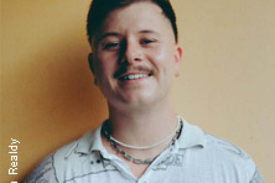 Ein junger Mann mit Schnurrbart lächelt in die Kamera.