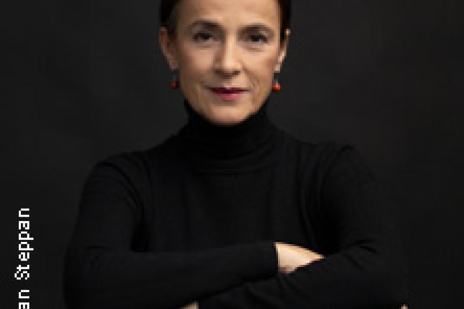 Eine Frau mit dunklen Haaren und einem schwarzen Rollkragenpullover sitzt auf einem Stuhl.