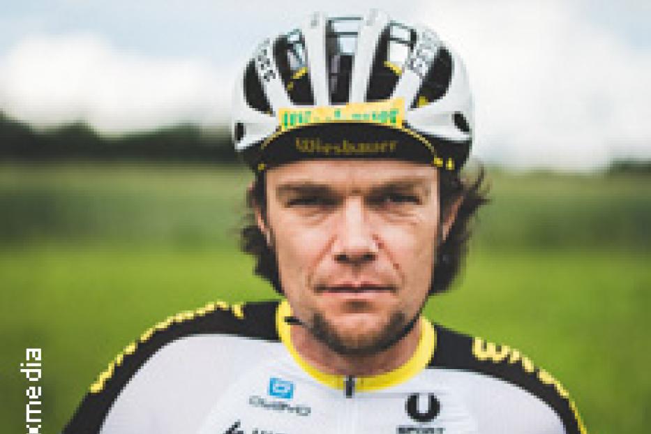 Ein Radfahrer in Sportkleidung und Helm steht vor einer grünen Landschaft.