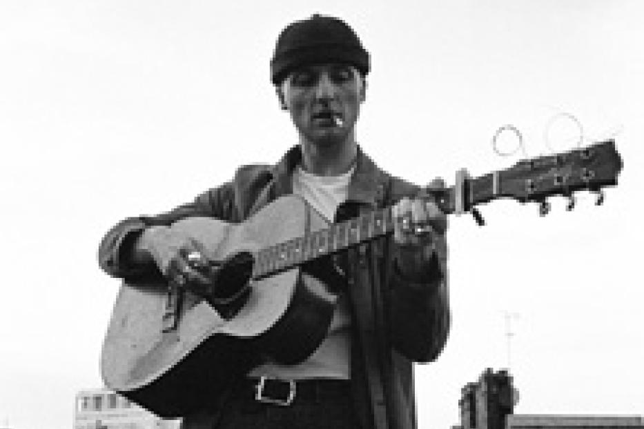 Ein Mann mit Mütze und Mantel spielt Gitarre und raucht eine Zigarette.