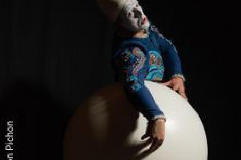 Ein Clown mit weißem Gesicht und blauem Kostüm sitzt auf einer großen, weißen Kugel.