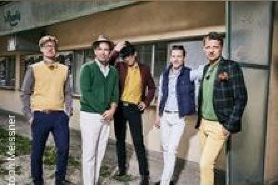 Fünf Männer posieren in farbenfrohen Outfits vor einem Gebäude.