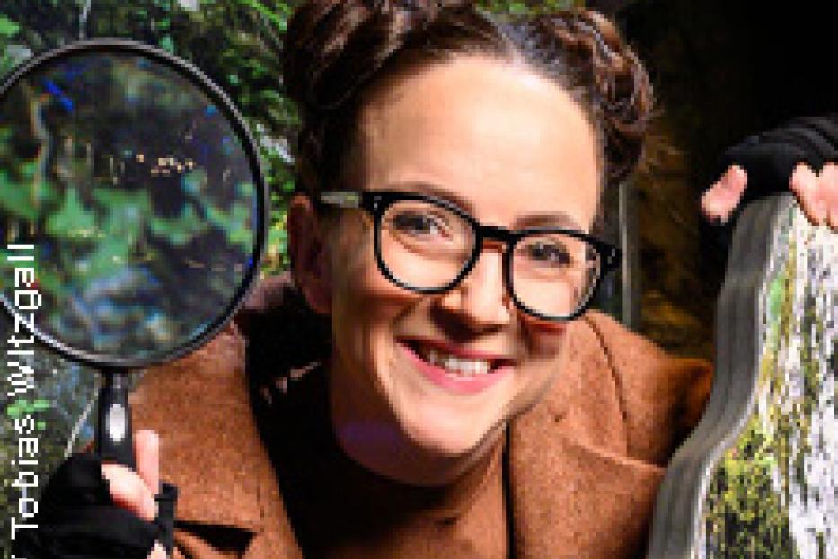 Eine lächelnde Frau mit Brille und Dutt hält eine Lupe vor einem grünen Hintergrund.