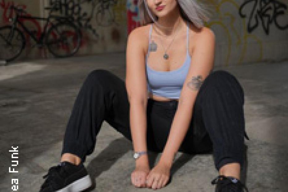 Eine junge Frau mit hellblondem Haar sitzt vor einer Graffitiwand.