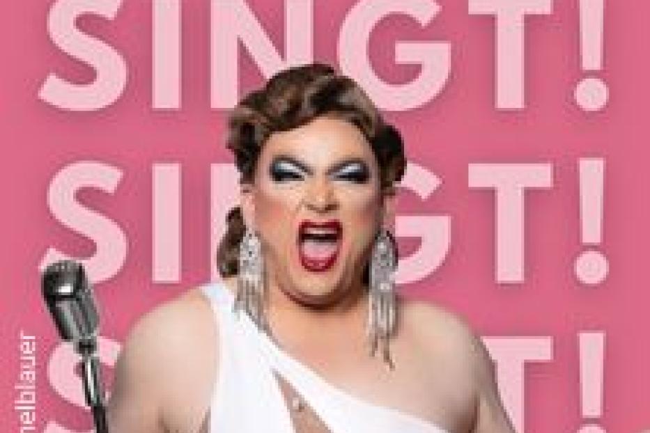 Eine Dragqueen mit Mikrofon singt vor einem pinkfarbenen Hintergrund mit dem Wort „Singt!“.