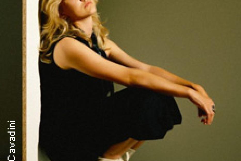 Eine Frau in einem schwarzen Kleid und weißen Stiefeln kauert vor einer Wand.