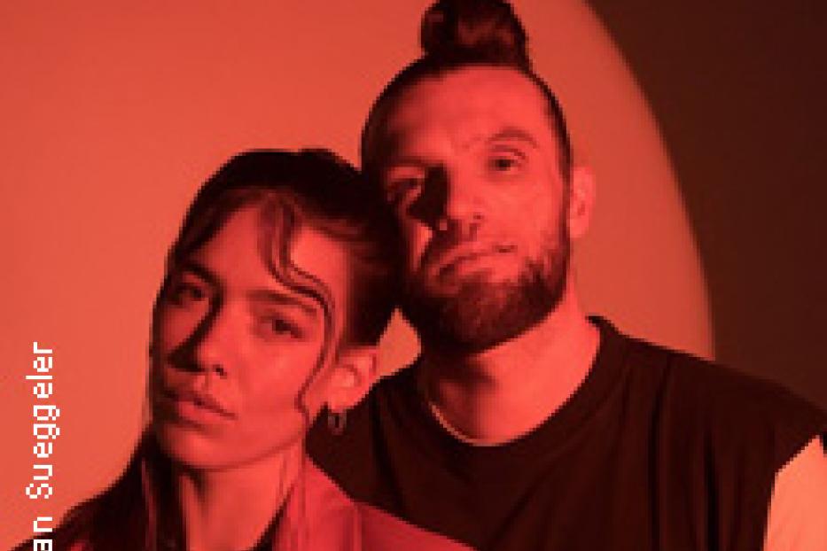 Ein Mann mit Dutt und eine Frau mit Blazer posieren vor einem roten Hintergrund.