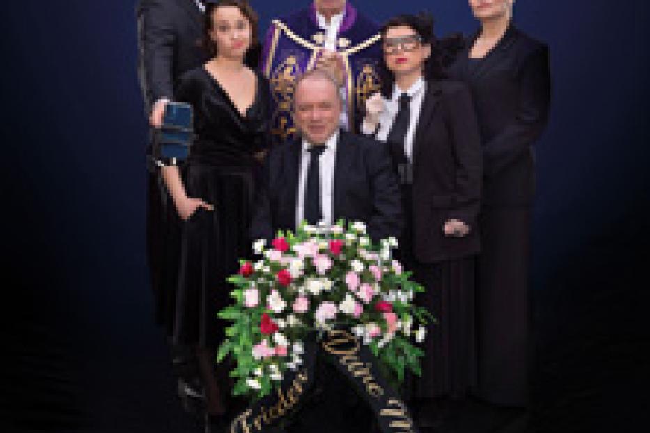 Eine Gruppe von Personen in Trauerkleidung steht um einen Kranz mit der Aufschrift „Ruhe in Frieden, Deine Mutter Mitarbeiter Team“.