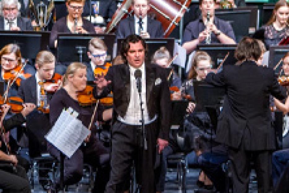 Ein Sänger steht vor einem Orchester und singt in ein Mikrofon.