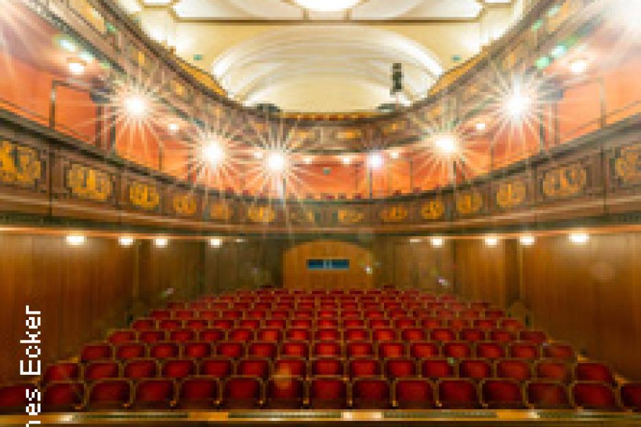 Innenansicht eines Theaters mit roten Sitzen und kunstvoller Holzvertäfelung.