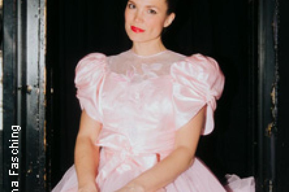Eine Frau in einem rosa Kleid sitzt vor einem dunklen Hintergrund.