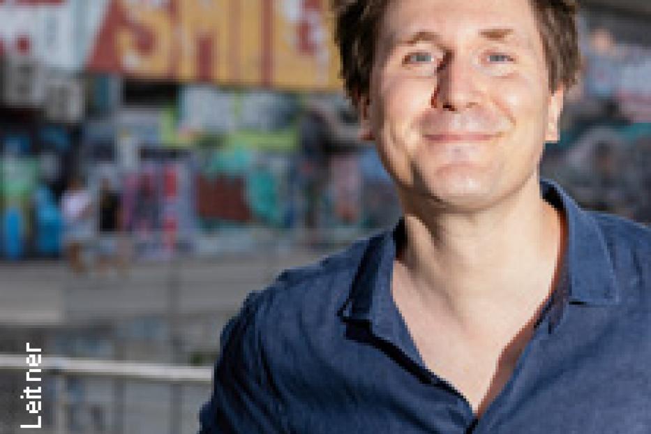 Ein Mann mit blauem Hemd lächelt vor einer Wand mit Graffiti.