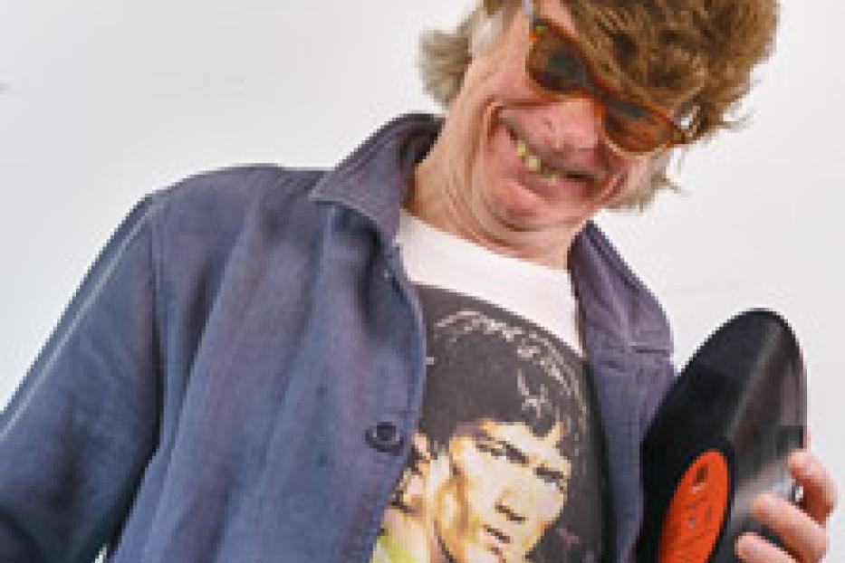 Ein Mann mit Perücke und Sonnenbrille lächelt und hält eine Schallplatte.