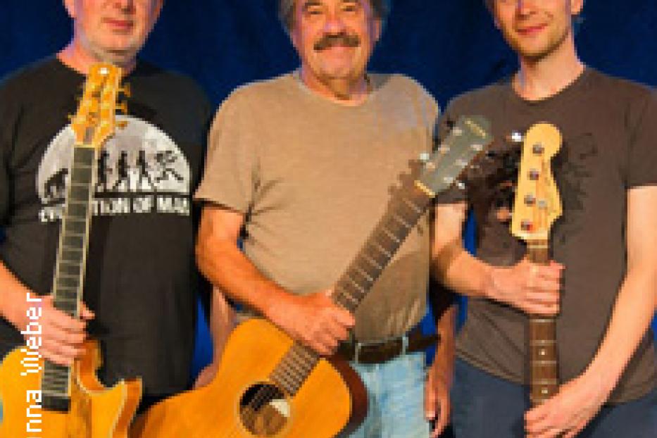 Drei Männer stehen mit Gitarren vor einem blauen Hintergrund.