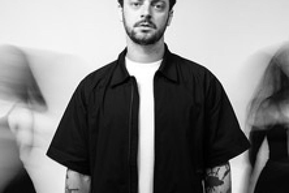 Ein Mann mit Bart und dunklem Hemd steht vor einem hellen Hintergrund.